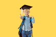 Child boy primary school student in a graduate cap, glasses and with a backpack on a yellow background. Shows ok sign with his finger. Knowledge day and back to school. Copy space