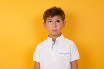 little cute Caucasian boy kid wearing white t-shirt against yellow wall crosses eyes, puts lips, makes grimace with awkward expression has fun alone, plays fool.