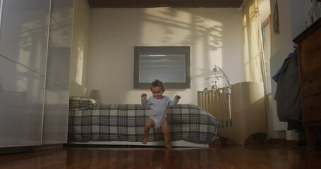 Wall Mural - Cinematic shot of happy smiling barefoot little toddler baby boy wearing light blue body is making his first steps on wooden floor at home. Concept: life, childhood, authenticity, parenthood, home.