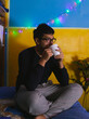 © JuanCarlos - hombre joven sentado en la orilla de su cama con luces de fondo de navidad realizando distintas acciones enfrente de una laptop mientras sostiene una taza de café durante la pandemia de Covid-19