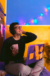 © JuanCarlos - hombre joven sentado en la orilla de su cama con luces de fondo de navidad realizando distintas acciones enfrente de una laptop mientras sostiene una taza de café durante la pandemia de Covid-19