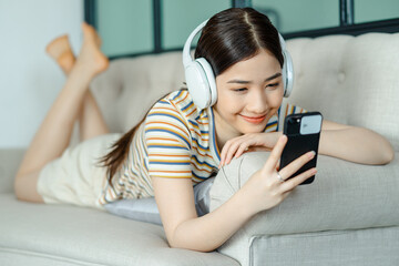 beautiful asian woman lying on sofa and listening to music while using phone