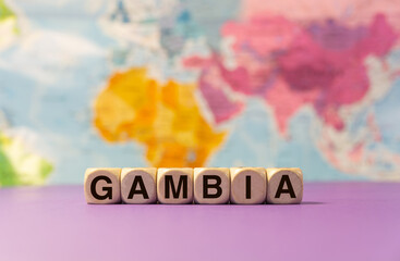 The word Gambia written with wooden dices in front of a purple background and a geographic map