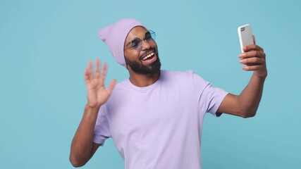 Wall Mural - Funny african american man in violet t-shirt hat isolated on blue background studio. People lifestyle concept. Talk on mobile phone making video call greeting showing victory sign Ok gesture thumb up