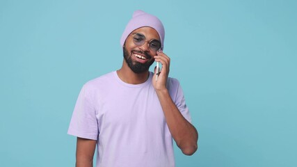 Wall Mural - Joyful excited young african american man 20s in violet t-shirt hat isolated on blue turquoise background studio. People lifestyle concept. Talking on mobile cell phone doing winner gesture screaming