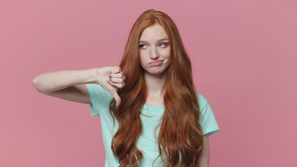 Wall Mural - Displeased ginger redhead young woman in casual blue turquoise t-shirt posing isolated on pink background studio. People lifestyle concept. Looking camera say no with finger gesture showing thumb down