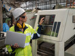 © panyajampatong - Confident young female mechanical engineer in a protective uniform, helmet, and eyeglasses use a Laptop computer to calibrate the CNC Machinery while working at the plant.