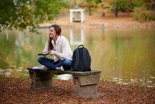 Beautiful Young College Student Sits Alone On Campus In Fall Leaves Near Small Pond - Holding Tablet Computer And Laughing