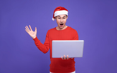 Santa asian man in red casual attire holding laptop and looking at screen excited face and surprise hand on purple background.Happy new year concept.