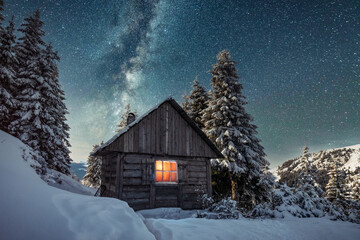 Fantastic winter landscape with wooden house in snowy mountains. Starry sky with Milky Way and snow covered hut. Christmas holiday and winter vacations concept