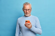 © WHstudio Leushin N - Photo of satisfied bearded man focused in smart phone surfs internet sends text messages in social networks uses modern technologies wears casual blue jumper poses indoor. Senior freelancer.