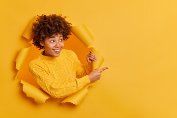 Wall Mural - Try this! Cheerful pleasant looking young Afro American woman points away on copy blank space dressed in casual jumper breaks through paper wall gives recommendation asks to use good opportunity