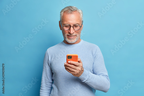 Cuadros en Lienzo Photo of satisfied bearded man focused in smart phone surfs internet sends text messages in social networks uses modern technologies wears casual blue jumper poses indoor