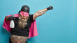 © WHstudio Leushin N - Shocked bearded superhero with cleaning agent stretches arm ready to clean everything on his way wears pink cloak helmet and mask has clothespins stuck in thick beard isolated over blue background