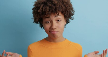 Wall Mural - Indecisive brunette woman shrugs shoulders and raises palms in bewilderment cannot make choice stands doubtful indoor wears casual orange jumper being questioned isolated over blue background