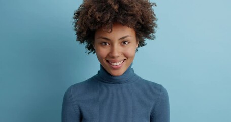 Wall Mural - Glad smiling young Afro American woman with perfect white teeth has positive expression looks happily at camera dressed in blue poloneck chuckles as hears something good or funny poses indoor.