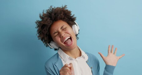 Wall Mural - Upbeat dark skinned woman enjoys favorite playlist listens music in headphones raises arms and dances carefree has fun feels every bit isolated over blue background sings song makes microphone