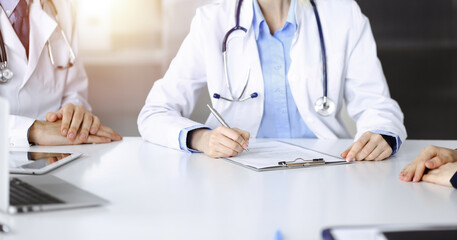 Unknown woman-doctor with male colleague are consulting patient woman while sitting at the desk in sunny clinic, close-up. Covid 2019