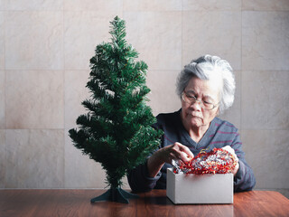 Elderly Asian woman decorating the Christmas tree while sitting in a living room at home. Space for text. Concept of aged people and Christmas and happy new year festival