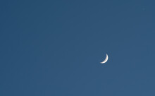 A Slim Sliver Of The Silvery Moon Set On An Expanse Of A Partly Cloudy Blue Sky.
