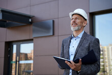 Monitoring The Process. Confident Middle Aged Architect, Engineer Manager Wearing Helmet Looking Away While Standing Outdoors And Holding A Blueprint