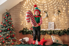 Little Cheerful Boy In An Elf Hat Jumping On The Bed With An Inflatable Cane In His Hands