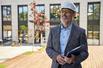 Providing urban design. Successful middle aged architect, engineer manager wearing helmet smiling, standing outdoors and holding a blueprint in his hands while planning new project