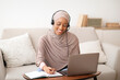 © Prostock-studio - Happy black woman in hijab wearing headphones, watching webinar on laptop, taking notes at home