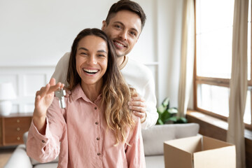 Happy spouses portrait, people standing in living room on relocation day smiling looking at camera, husband hug wife holding keys to the new house. Moving day, bank lending for young family concept