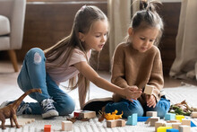Two Little 6s 7s Sisters Pretty Daughters Spend Free Playtime Seated On Carpet Warm Floor In Modern Living Room Play Together Wooden Brick And Dinosaurs Toys Set. Friendship, Fun And Leisure Concept