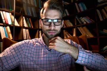 Serious young business man looking at camera or computer webcam working late elearning online video conference calling, or watching webinar at home office in night lamp light. web cam view. Headshot.