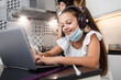 © Dusko - Young little girl having e-learning session during Covid-19 pandemic crisis lockdown or quarantine. Busy mother working in kitchen and preparing lunch in the background.