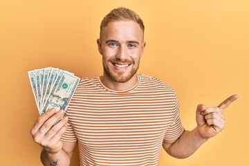 Wall Mural - Young caucasian man holding dollars smiling happy pointing with hand and finger to the side