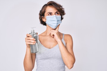 Wall Mural - Young hispanic woman wearing medical mask holding hand sanitizer gel serious face thinking about question with hand on chin, thoughtful about confusing idea