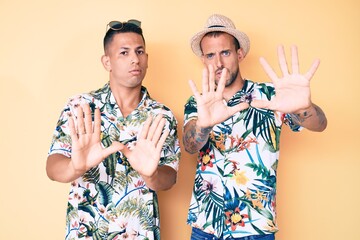 Wall Mural - Young gay couple of two men wearing summer hat and hawaiian shirt moving away hands palms showing refusal and denial with afraid and disgusting expression. stop and forbidden.