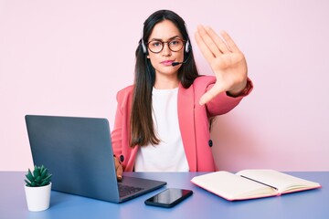 Poster - Young caucasian woman sitting at the desk wearing call center agent headset working using laptop with open hand doing stop sign with serious and confident expression, defense gesture