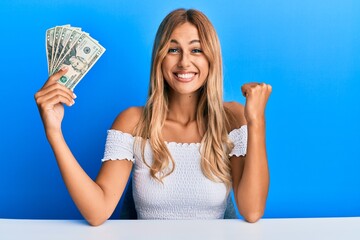 Wall Mural - Beautiful blonde young woman holding 20 dollars banknotes screaming proud, celebrating victory and success very excited with raised arm