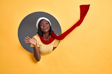 Portrait of friendly woman in santa hat and red scarf waving raised hand and saying hi to camera, enjoying christmas time. Indoor studio shot isolated in circle hole in orange background. Copy space.