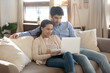 © fizkes - Happy bonding millennial couple in love having date at home embracing on soft comfy couch at living room spending time online using modern laptop pc device watching cute photo video at social networks