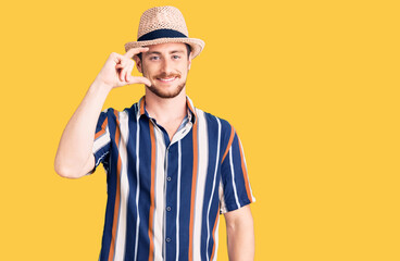 Young handsome caucasian man wearing summer hat smiling and confident gesturing with hand doing small size sign with fingers looking and the camera. measure concept.