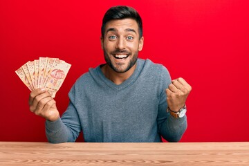 Handsome hispanic man holding mexican pesos screaming proud, celebrating victory and success very excited with raised arm