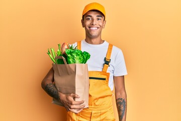 Young handsome african american man wearing courier uniform with groceries from supermarket looking positive and happy standing and smiling with a confident smile showing teeth