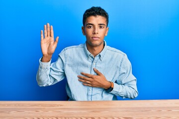 Sticker - Young handsome african american man wearing casual clothes sitting on the table swearing with hand on chest and open palm, making a loyalty promise oath