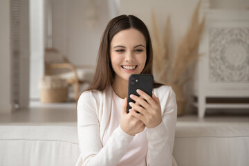 Leisure time. Cheerful happy young woman holding mobile telephone taking selfie on couch at home to post at social network profile, having pleasant talk by video link app, recording vlog using webcam