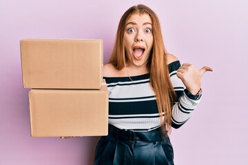 Young beautiful redhead woman holding delivery package pointing thumb up to the side smiling happy with open mouth