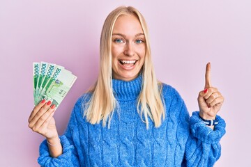 Sticker - Young blonde girl holding 10000 south korean won banknotes smiling with an idea or question pointing finger with happy face, number one