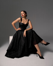 Elegant Lady In Black Shiny Ball Gown And Shoes Sitting At White Cibe With Crossed Legs And Posing Against Grey Background, Full Length Fashion Portrait. Sexy Female Model