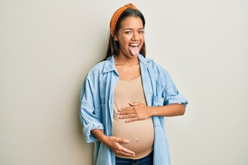 Sticker - Beautiful hispanic woman expecting a baby, touching pregnant belly sticking tongue out happy with funny expression. emotion concept.