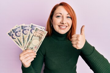 Sticker - Beautiful redhead woman holding japanese yen banknotes smiling happy and positive, thumb up doing excellent and approval sign