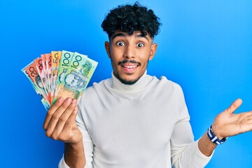 Young arab handsome man holding australian dollars celebrating achievement with happy smile and winner expression with raised hand